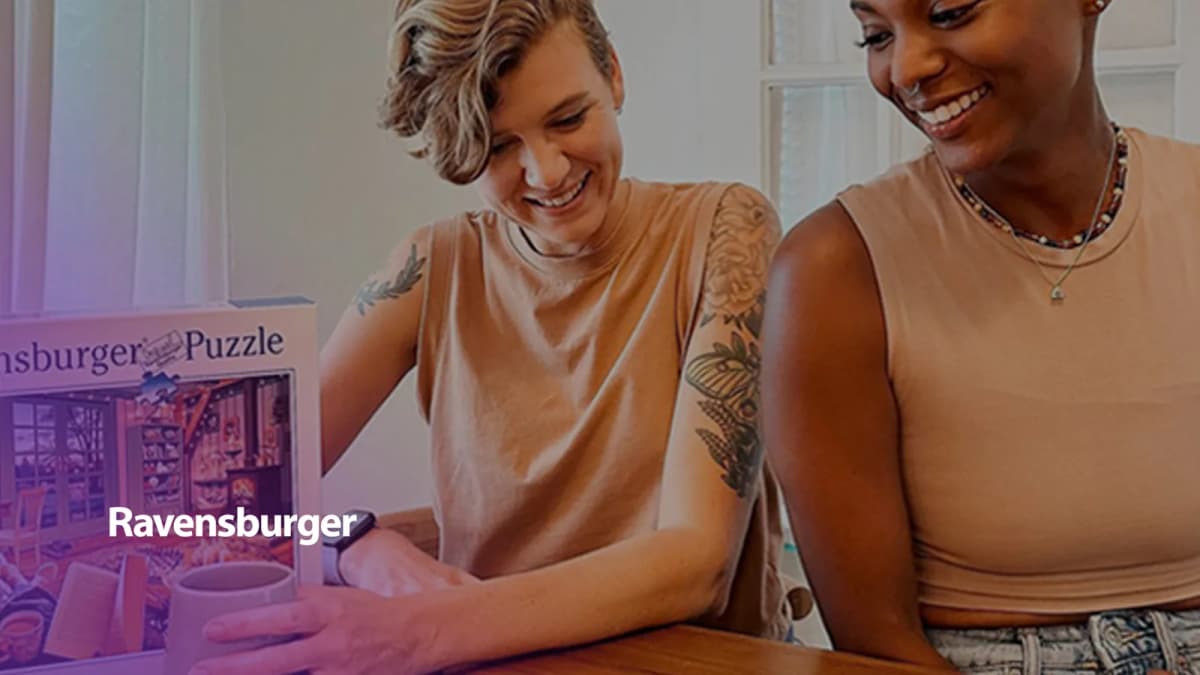 Two smiling people in tan tank tops look down at a table beside a Ravensburger "Puzzle" box with a cozy room scene