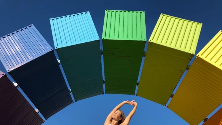 Woman stretching her arms overhead, eyes closed, beneath an arch of rainbow-colored shipping containers against blue sky