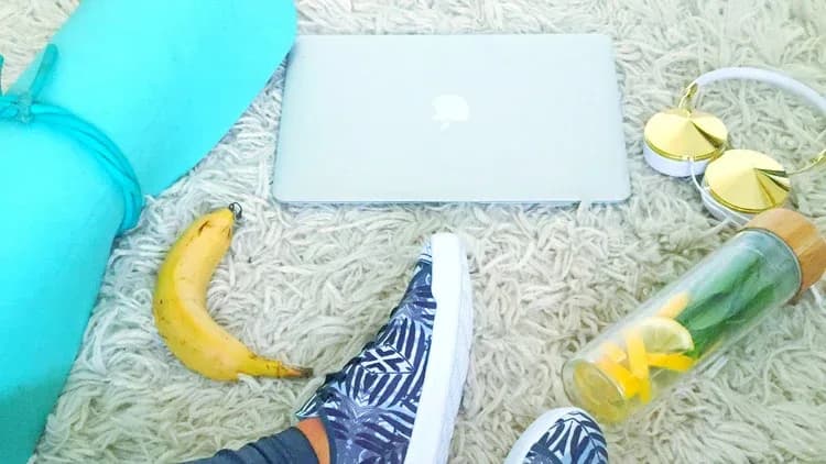 Feet in leaf-print sneakers on a shaggy rug near a yoga mat, banana, laptop, gold headphones and lemon water bottle
