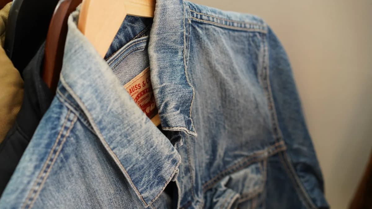 Close-up of a faded Levi's denim jacket on a wooden hanger, its collar label showing "AUSS & CO" and "CLOTHING"