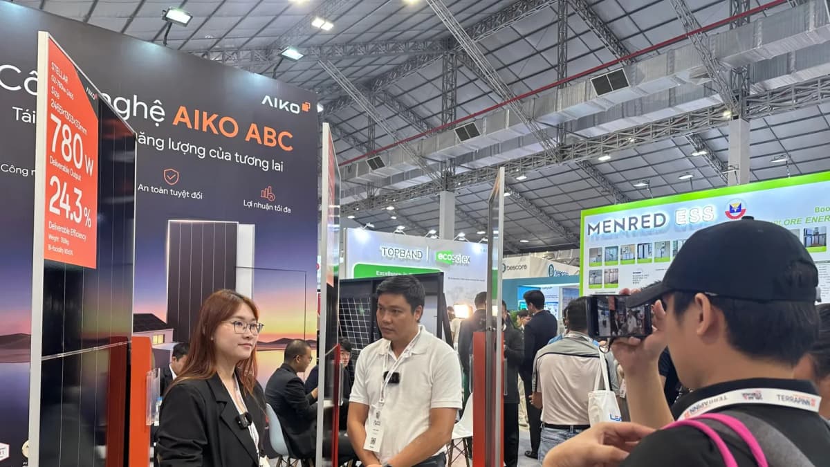 Man in a cap films a woman and man wearing mics at an AIKO Solar "AIKO ABC" panel booth at a busy trade show