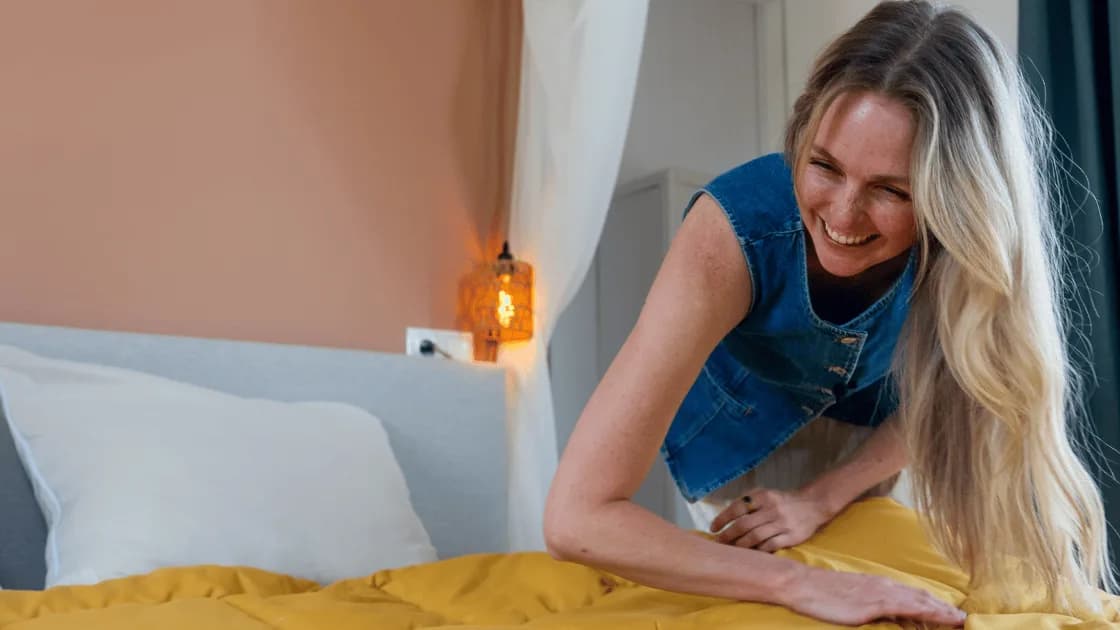 Smiling woman with long blond hair in a denim vest smooths a mustard-yellow duvet on a bed beside a glowing wall lamp
