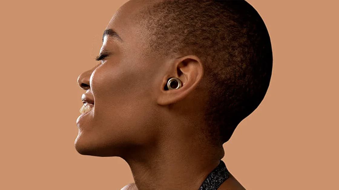 Smiling person with short cropped hair in profile wearing a gold ring-shaped Loop earplug, against a tan backdrop