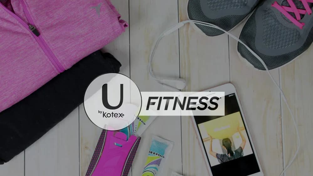 U by Kotex "FITNESS" logo over pink workout jacket, sneakers, earbuds, phone and wrapped pads on white wood floor