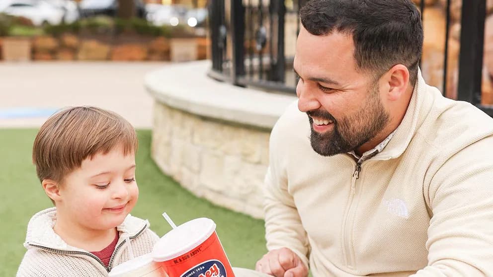 Bearded man smiles at a young boy beside a red Jersey Mike's Subs cup reading "BE A SUB ABOVE" outdoors