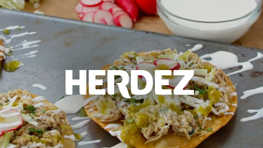 Herdez logo over shredded chicken tostadas with green salsa, crema, cabbage and radish on a baking sheet