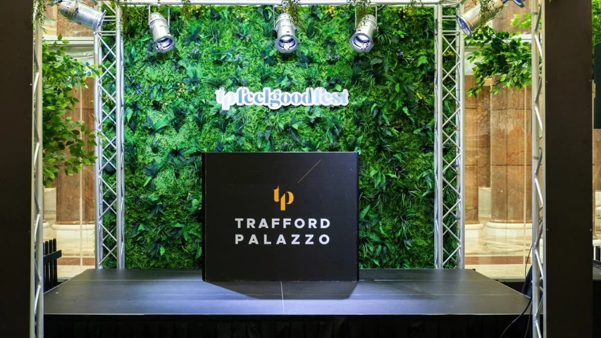 Empty stage with a "TRAFFORD PALAZZO" booth before a plant wall with a "tp feelgood fest" sign, lit by spotlights