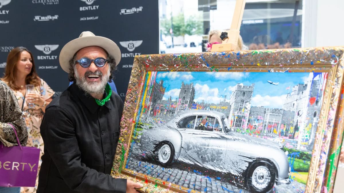 Bearded man in a hat and blue glasses holds a framed painting of a silver vintage car by a Clarendon Fine Art backdrop