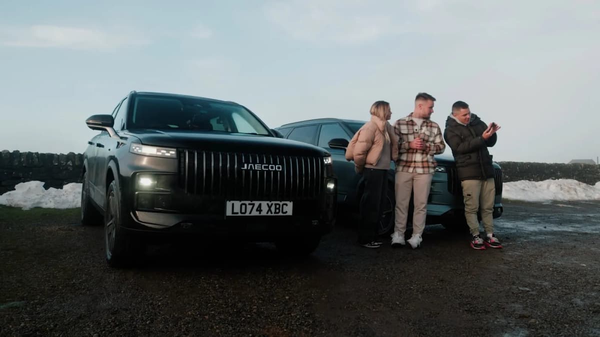 Black JAECOO SUV beside a second SUV where a woman and two men in winter coats look at a phone, snow by a stone wall